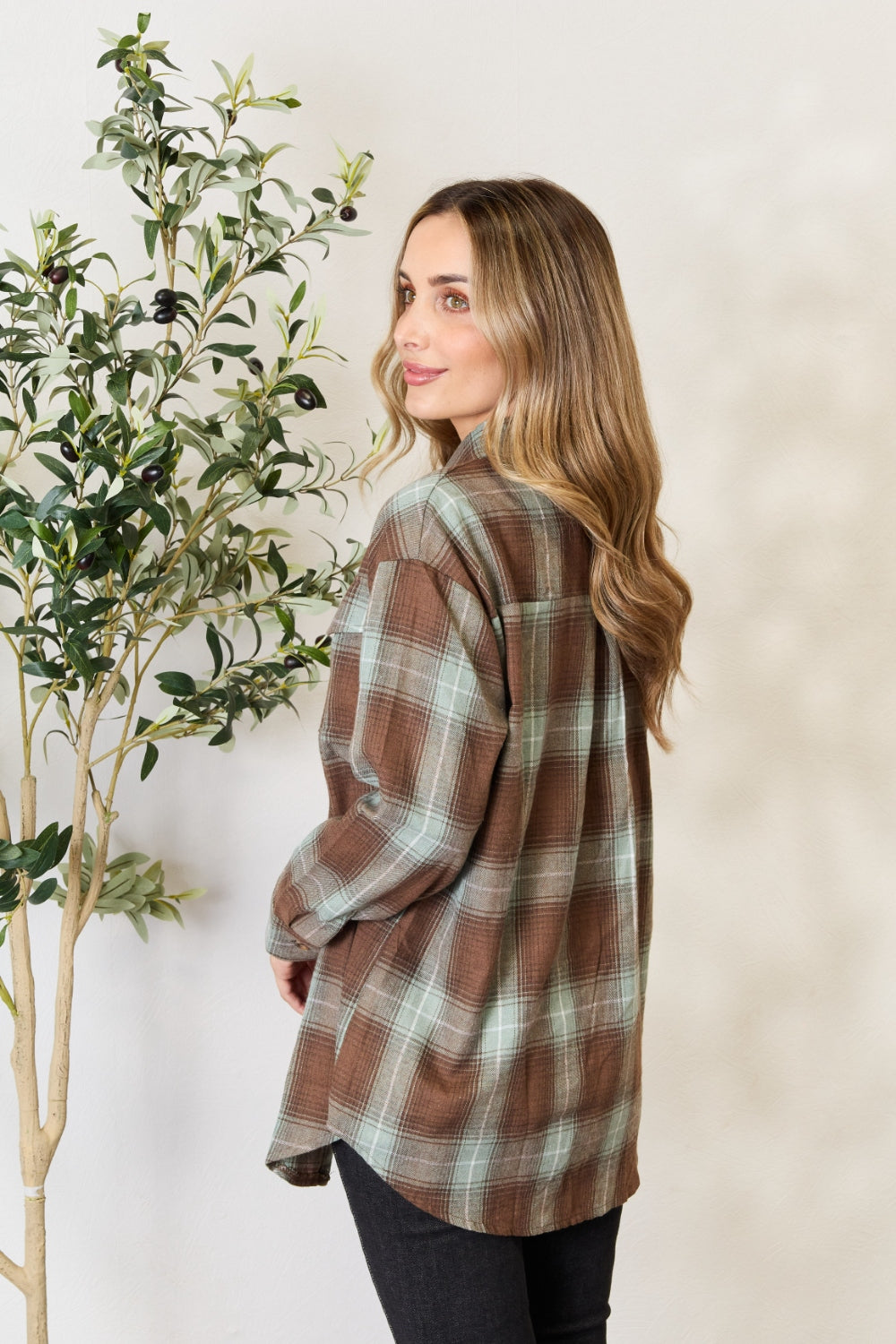 A woman stands in front of a light-colored wall and an olive tree, wearing the Mandy Plaid Dropped Shoulder Shirt paired with a white crop top and dark jeans. She smiles and holds her hair back with one hand.