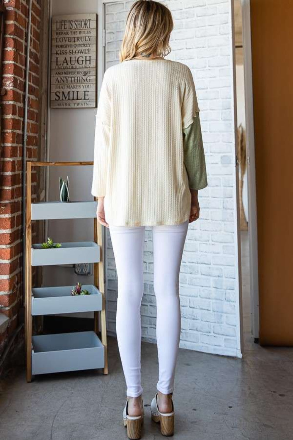 A person dressed in the Culture Code Color Block Round Neck Dropped Shoulder Blouse, featuring pastel shades of green, ivory, and lavender, stands in a room with a decorative sign on the brick wall.