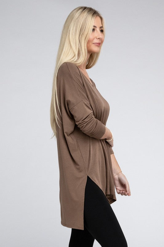 A woman with long, blonde hair is wearing a loose-fitting, brown Dolman Long Sleeve V-Neck Side Slit Hi-Low Hem Top and black pants. She poses with one hand on her hip and a neutral facial expression against a light grey background.