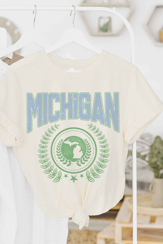 An individual dons a white MICHIGAN Graphic Tee, crafted from 100% cotton. The shirt prominently displays the word "Michigan" in blue, alongside an Earth graphic encircled by laurel leaves and topped with a star. Its unisex sizing guarantees a comfortable fit for all wearers.