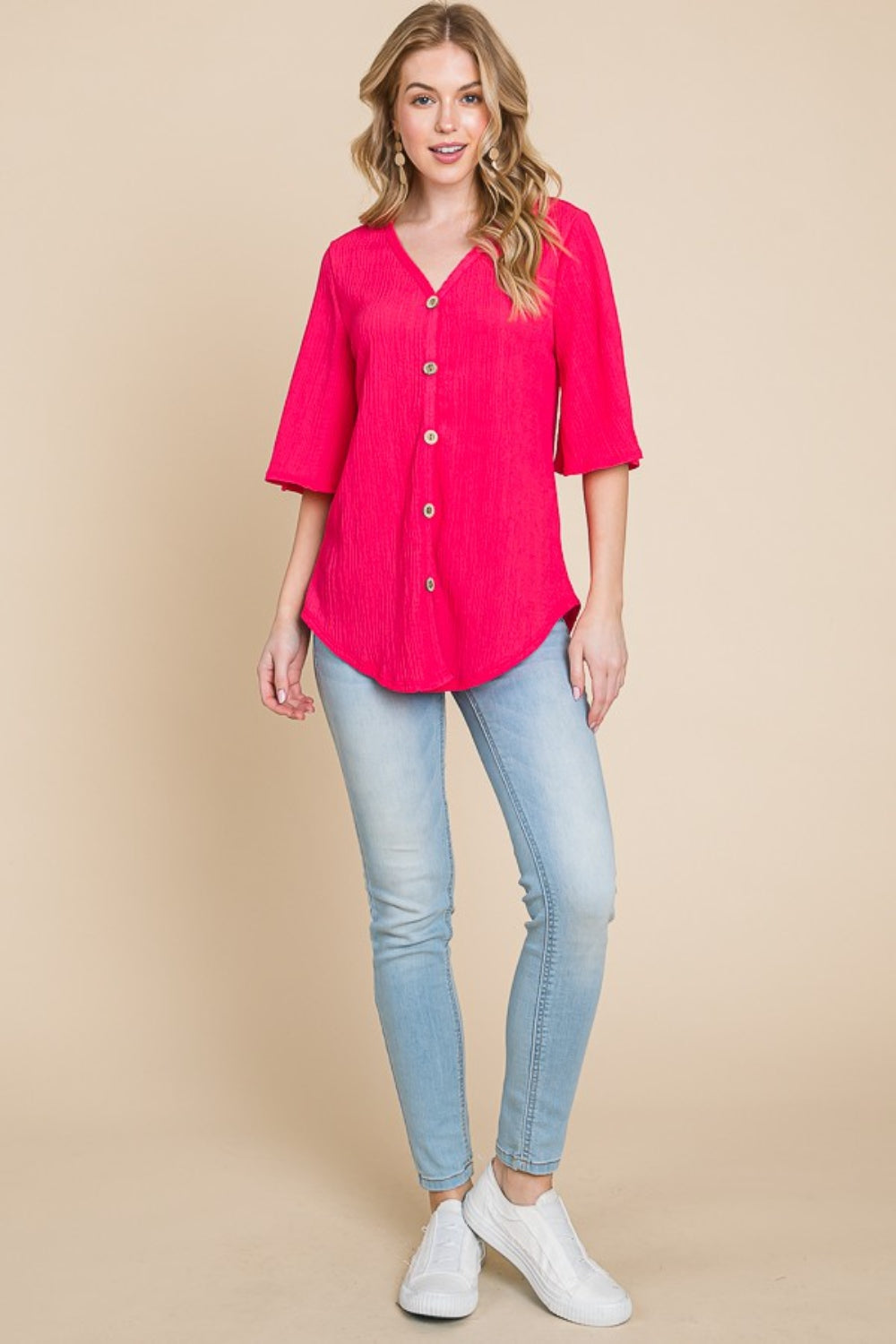 A person is wearing the BOMBOM Texture Decorative Button V-Neck Top, a chic design in bright pink featuring elegant details like short bell sleeves, and pairing it with blue jeans, stands against a beige background.