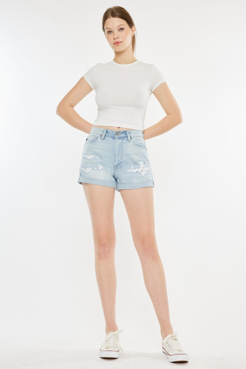 A person stands against a plain background, wearing a white short-sleeve crop top, Kancan High Rise Repaired Mom Denim Shorts in light blue, and white sneakers.