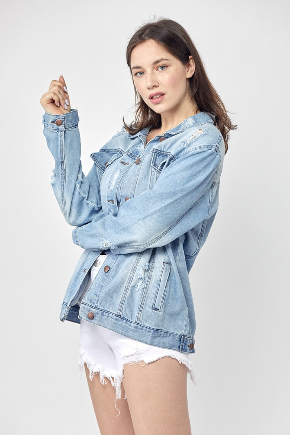 Dressed in the RISEN Full Size Distressed Long Sleeve Denim Jacket, a perfect wardrobe staple, this person effortlessly poses against a plain background while pairing it with a blue top and white shorts.