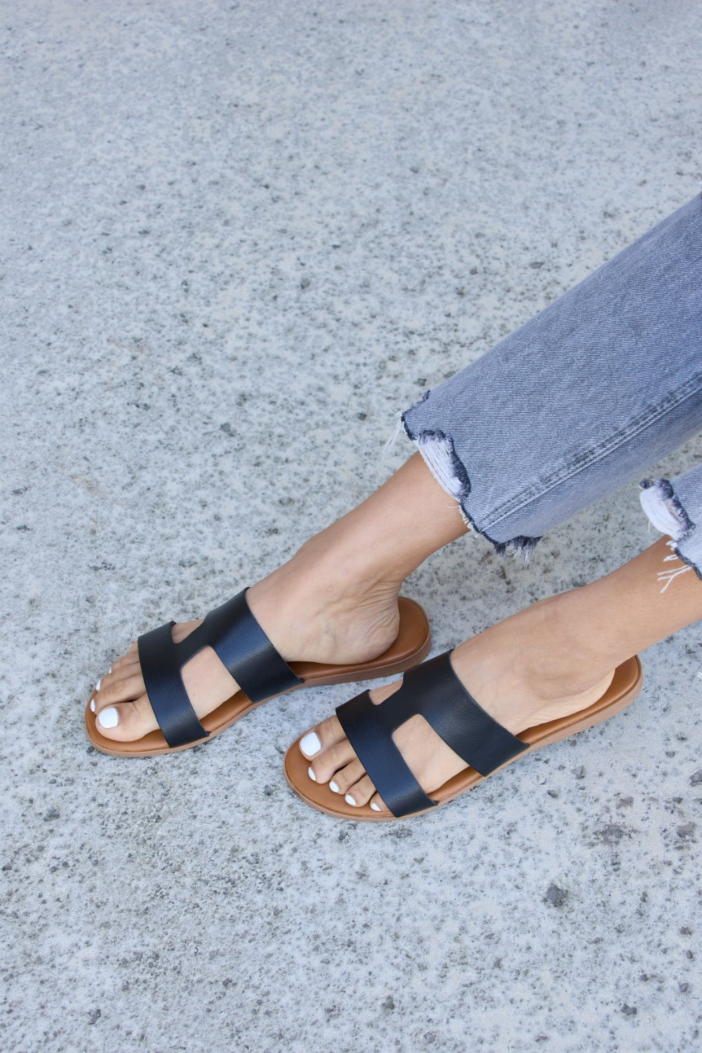 Someone wearing Forever Link Cutout Open Toe Flat Sandals and distressed jeans on a textured concrete surface.