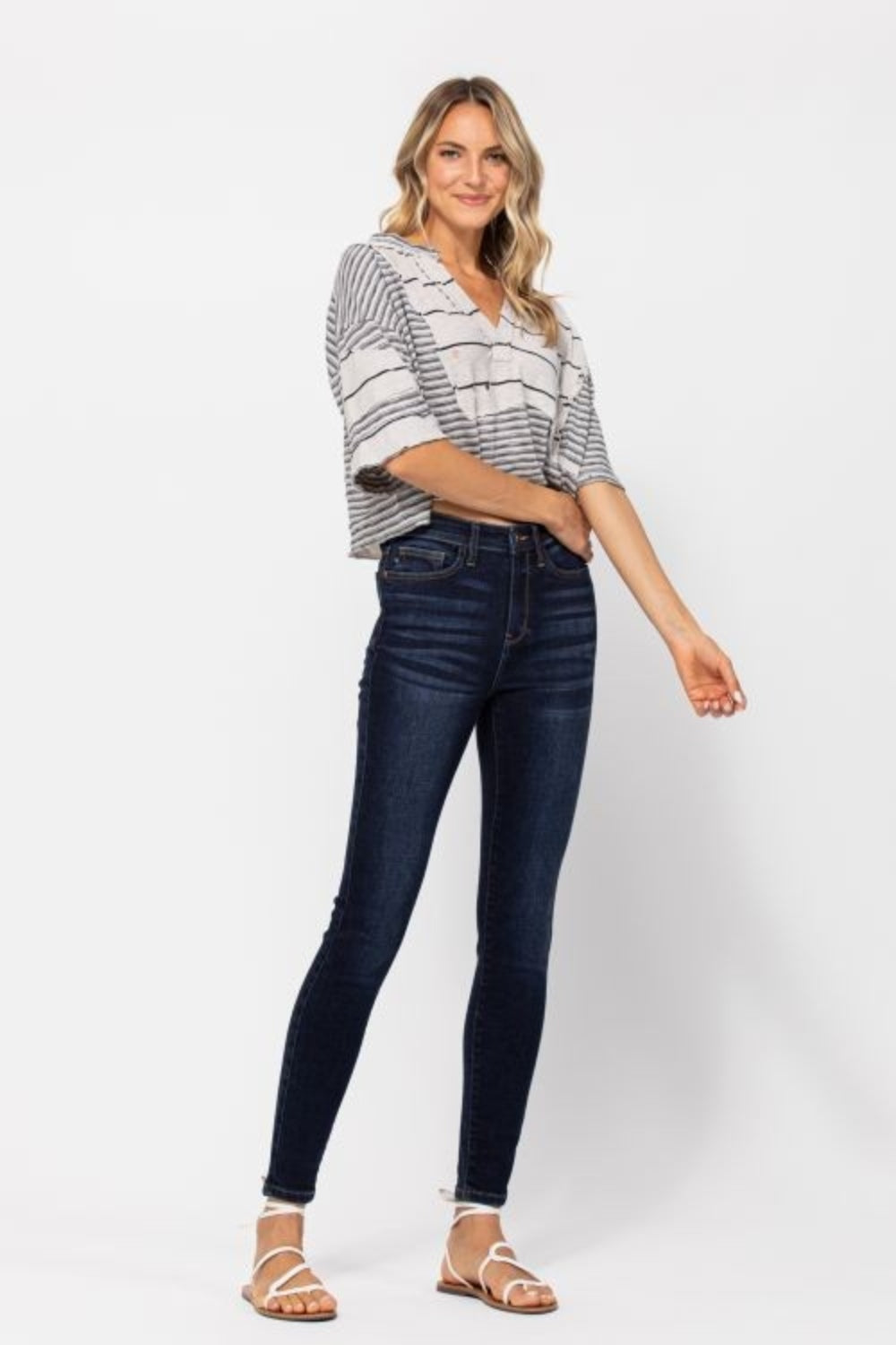 A person wearing versatile Judy Blue Full Size High Waist Handsand Skinny Jeans in dark blue and white strappy sandals is shown standing against a plain background.