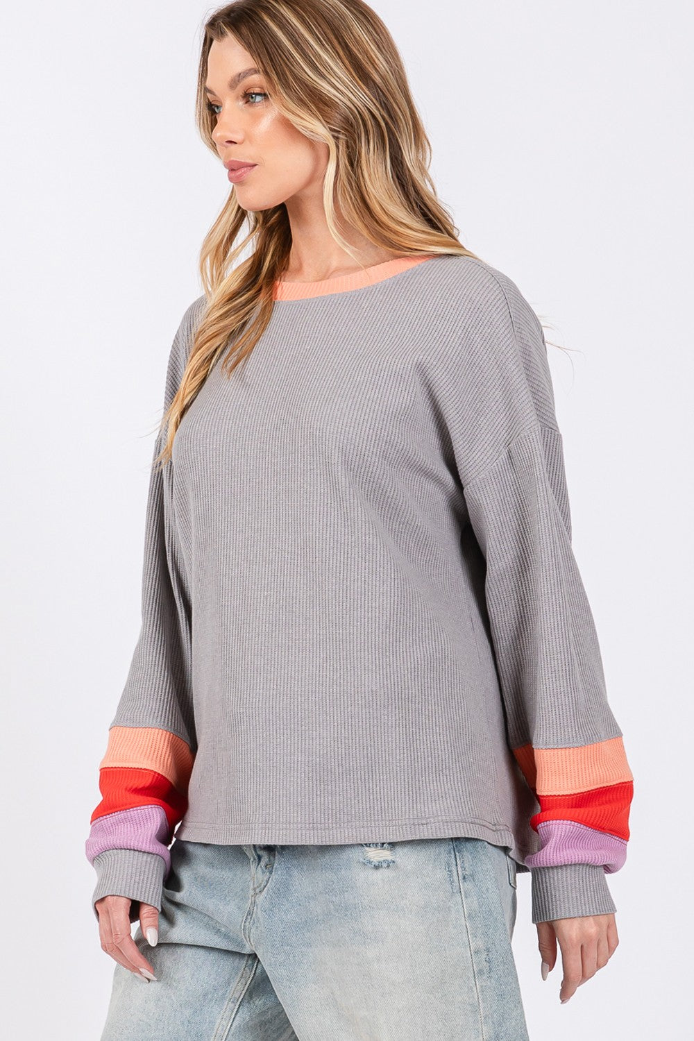 A woman wearing the SAGE + FIG Color Block Waffle Knit T-Shirt stands with one hand on her gray hat. The casual top, adorned with colorful block raglan sleeves, pairs effortlessly with denim shorts for a chic look.