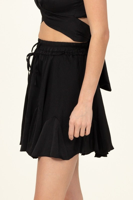 A person is partially visible in the image wearing the Not Your Girl Drawstring Ruffled Mini Skirt, featuring a bright pink flared silhouette with an elastic waistband and drawstring, paired with a matching top.