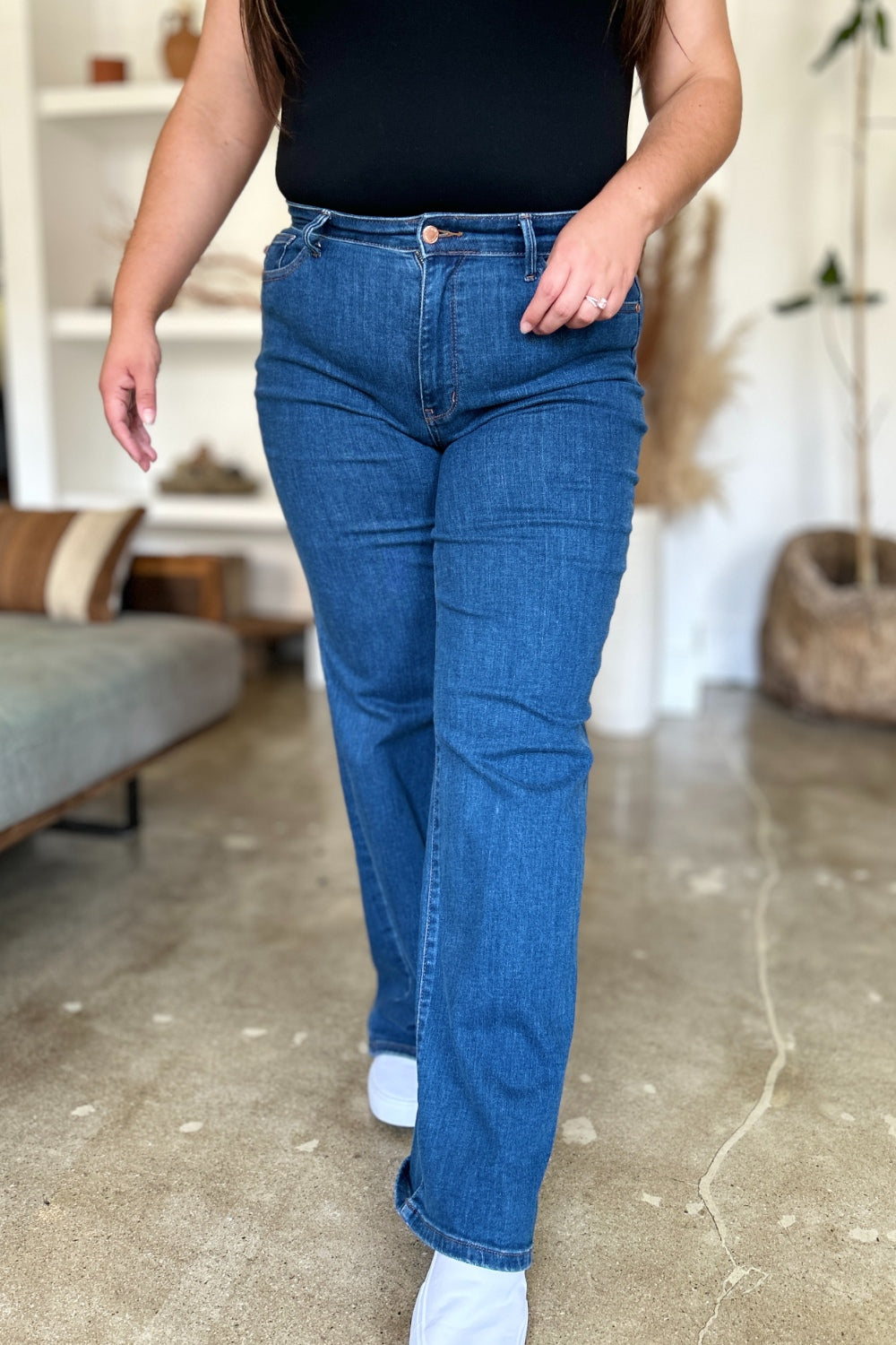 A person wearing Judy Blue Full Size High Rise Straight Jeans, a black top, and white shoes stands gracefully on a polished concrete floor.