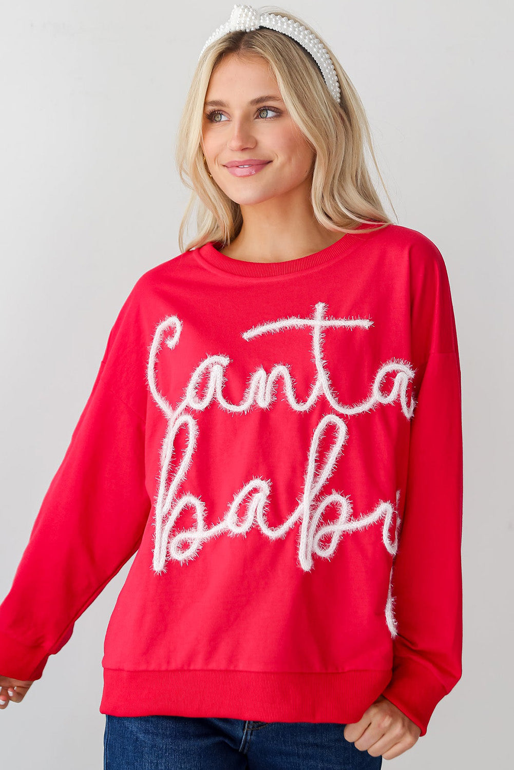 A person with long blonde hair is seen wearing the Fiery Red Christmas Santa Baby Tinsel Graphic Sweatshirt and blue jeans, facing away from the camera against a plain background.