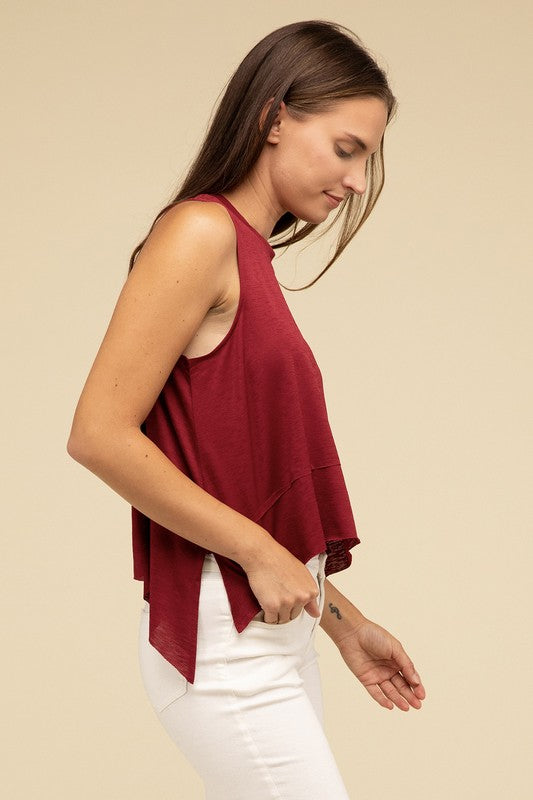 Wearing the Shark Bite Side Slit Short Sleeveless Top in maroon paired with white pants, a model is set against a beige background, exemplifying an effortlessly chic and casual wardrobe.