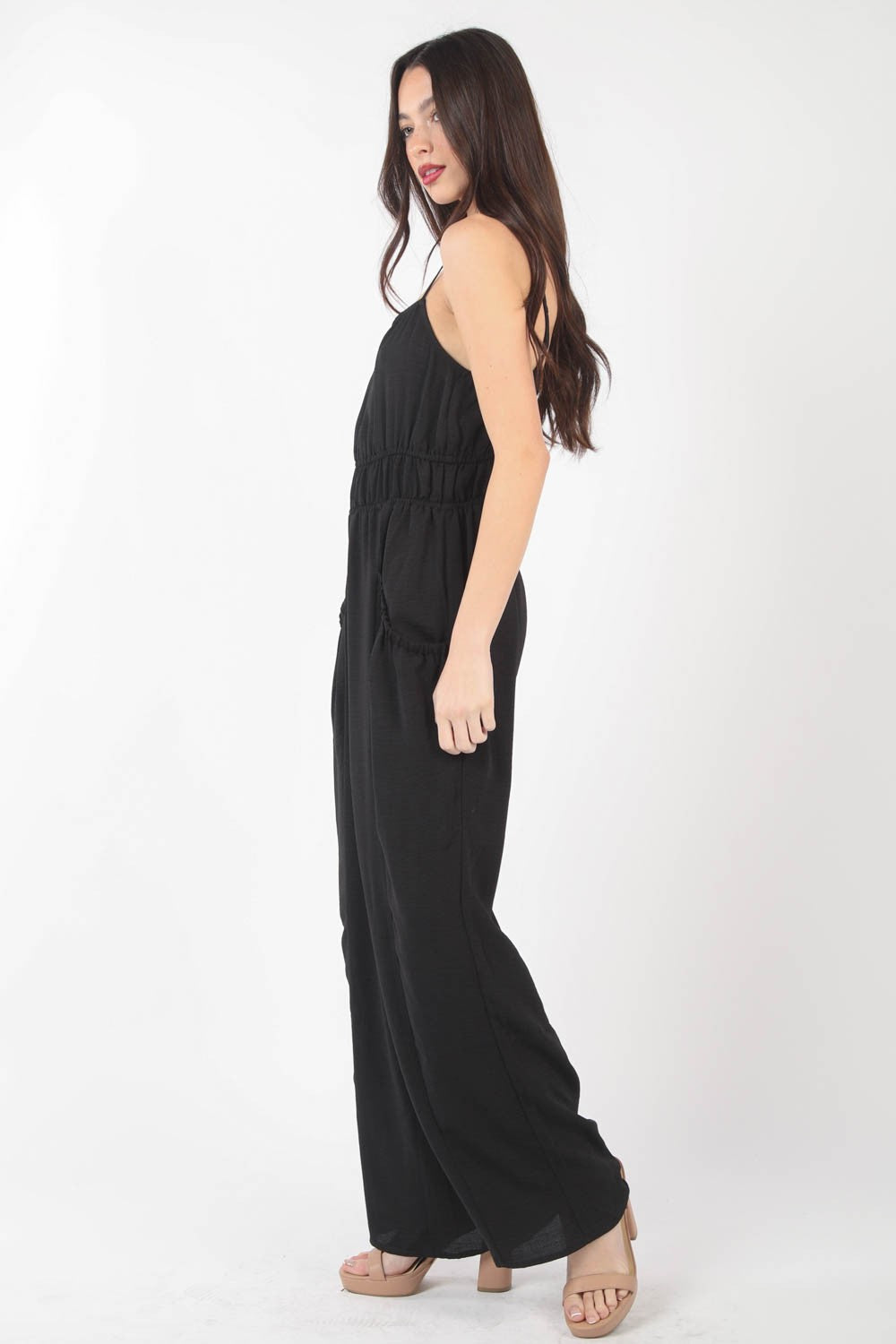 A woman stands against a plain backdrop wearing the VERY J Pintuck Detail Woven Sleeveless Jumpsuit. The black jumpsuit has thin straps, wide-leg pants, and is crafted from woven fabric with subtle pintuck details. She has her hands in her pockets and is looking at the camera.