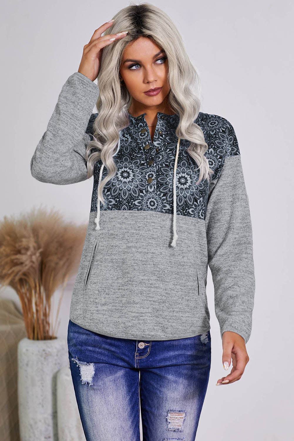 A person with long blonde hair is wearing a Gray Tribal Print Vintage Pocket Hoodie. They are turned sideways, and pampas grass in a vase is visible in the background.