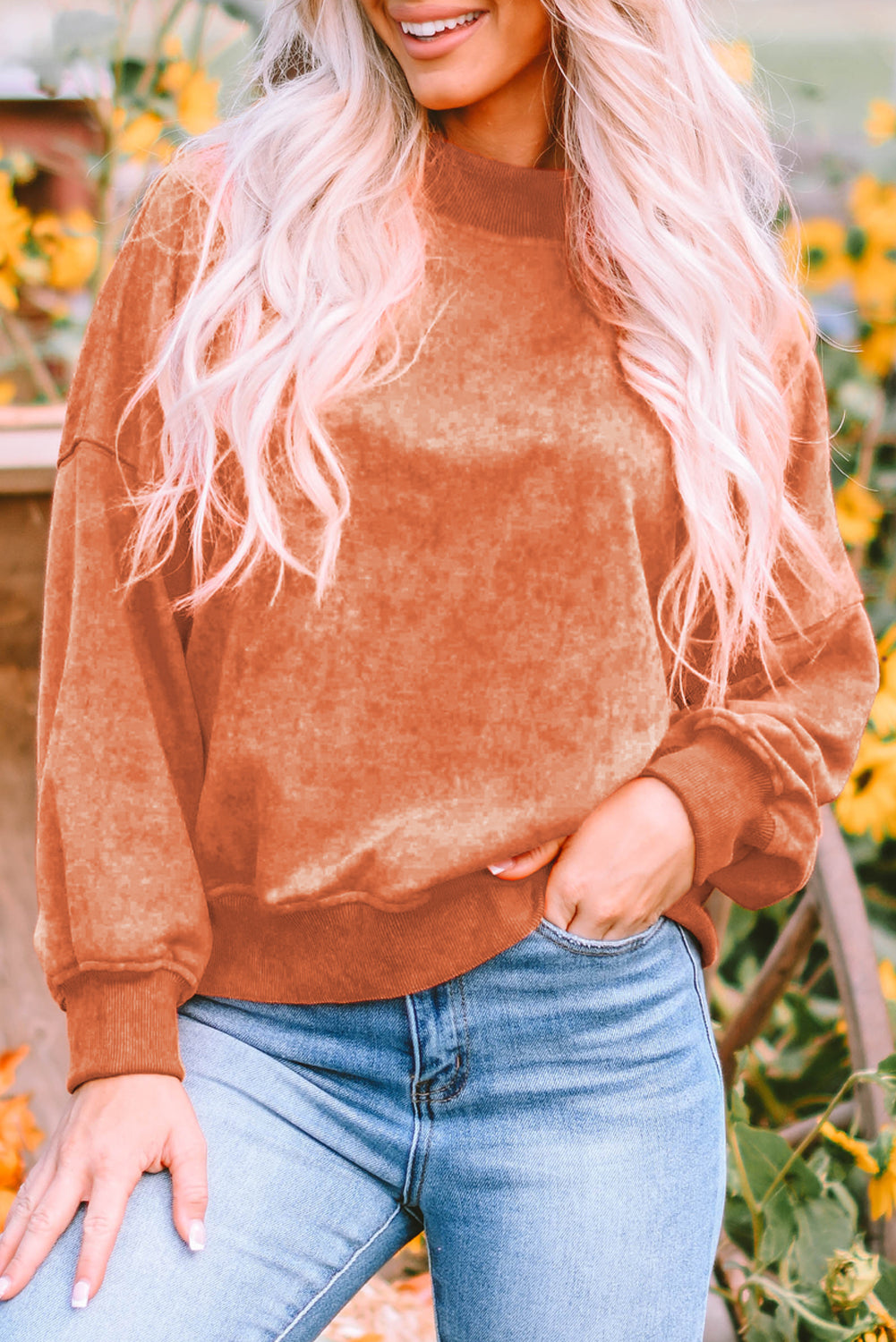 A person with long blonde hair wearing an Orange Drop Shoulder Crew Neck Pullover Sweatshirt and blue jeans stands with their back to the camera in a sunflower field.