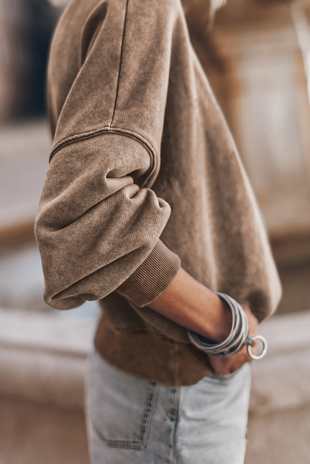 A person is seen wearing a brown Drop Shoulder Crew Neck Pullover Sweatshirt with rolled-up sleeves, paired with light blue jeans and standing with one hand in their pocket. Multiple bracelets are visible on their wrist while the background appears blurred.