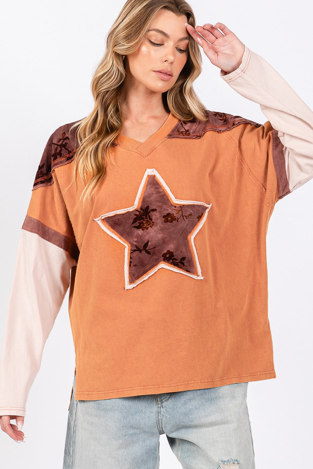A person wearing a beige hat, a SAGE + FIG Star Patch Long Sleeve Color Block T-Shirt in orange, and light blue jeans stands against a plain background.