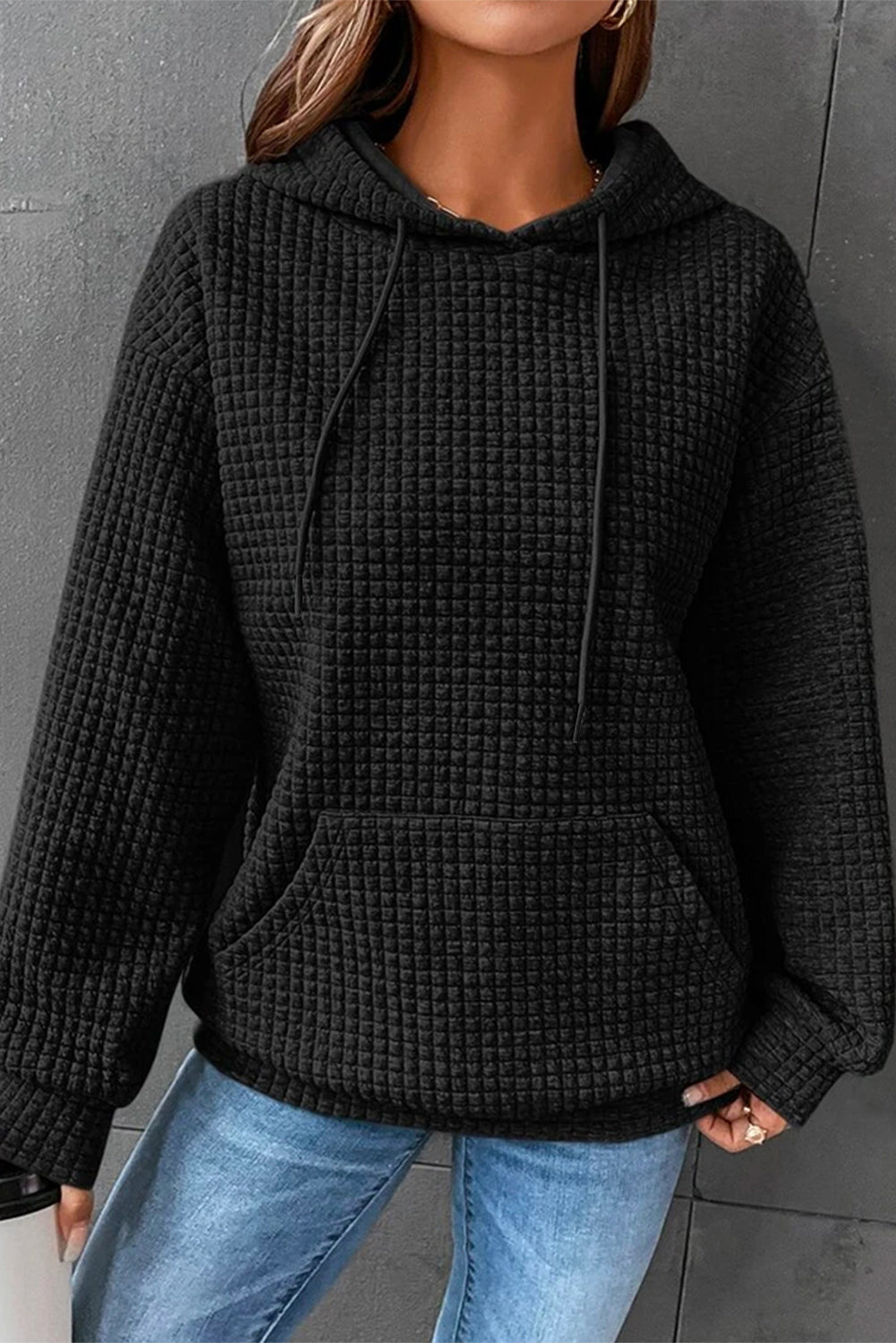 A person wearing a Black Quilted Kangaroo Pocket Drawstring Hoodie and blue jeans, with their left hand in the hoodie pocket and holding a black item in their right hand.