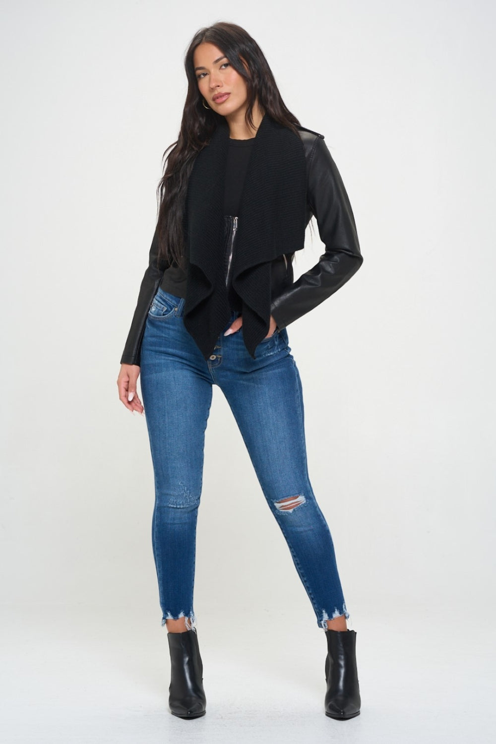 A woman with long, dark hair wearing a black top, a black shawl over the Coalition LA Knit Collared Faux Leather Crop Jacket, and blue jeans stands against a plain white background.