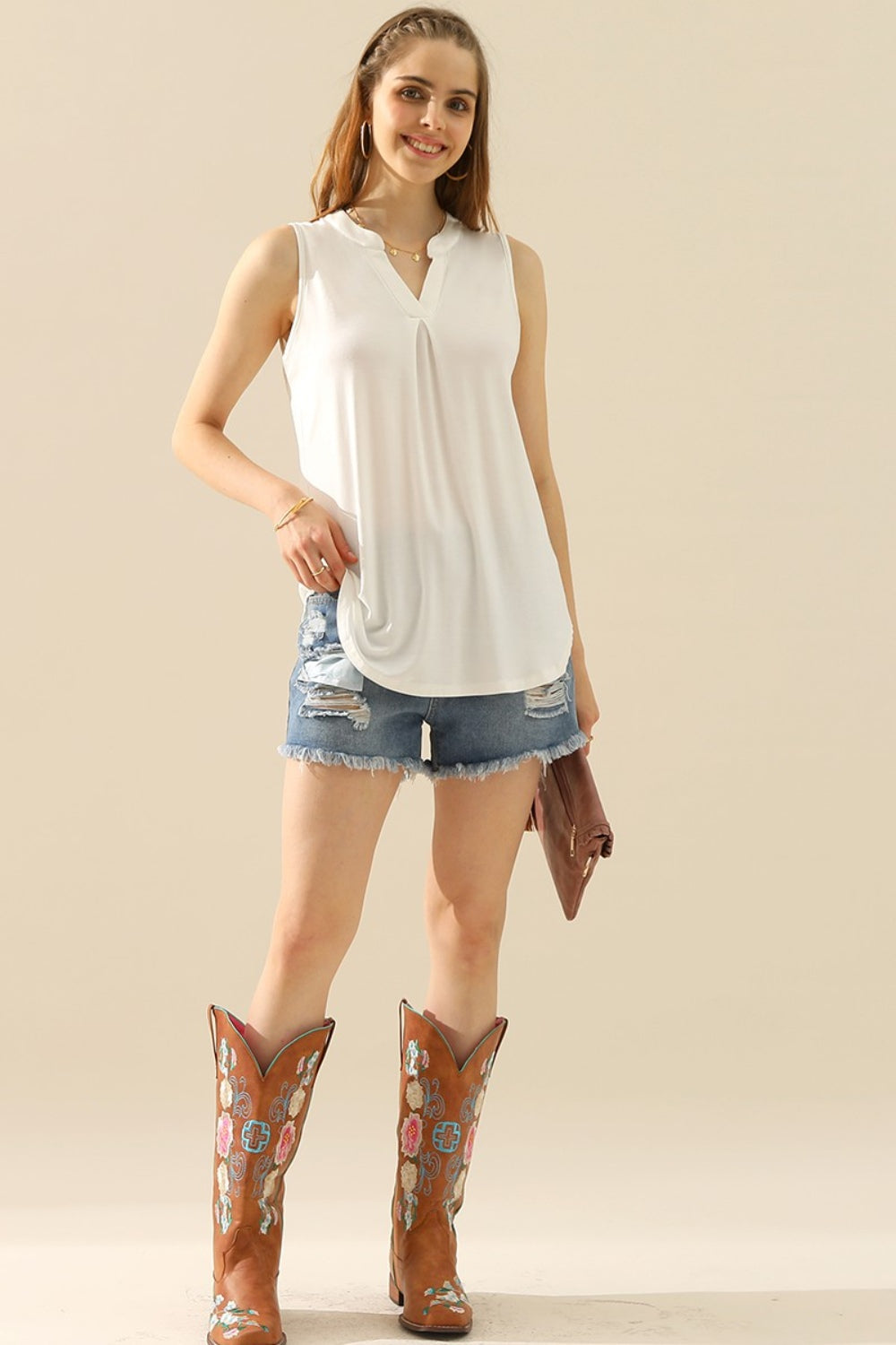 A person is standing while holding a brown clutch, wearing a Ninexis Full Size Notched Sleeveless Top in black, denim shorts, and brown cowboy boots against a neutral background.