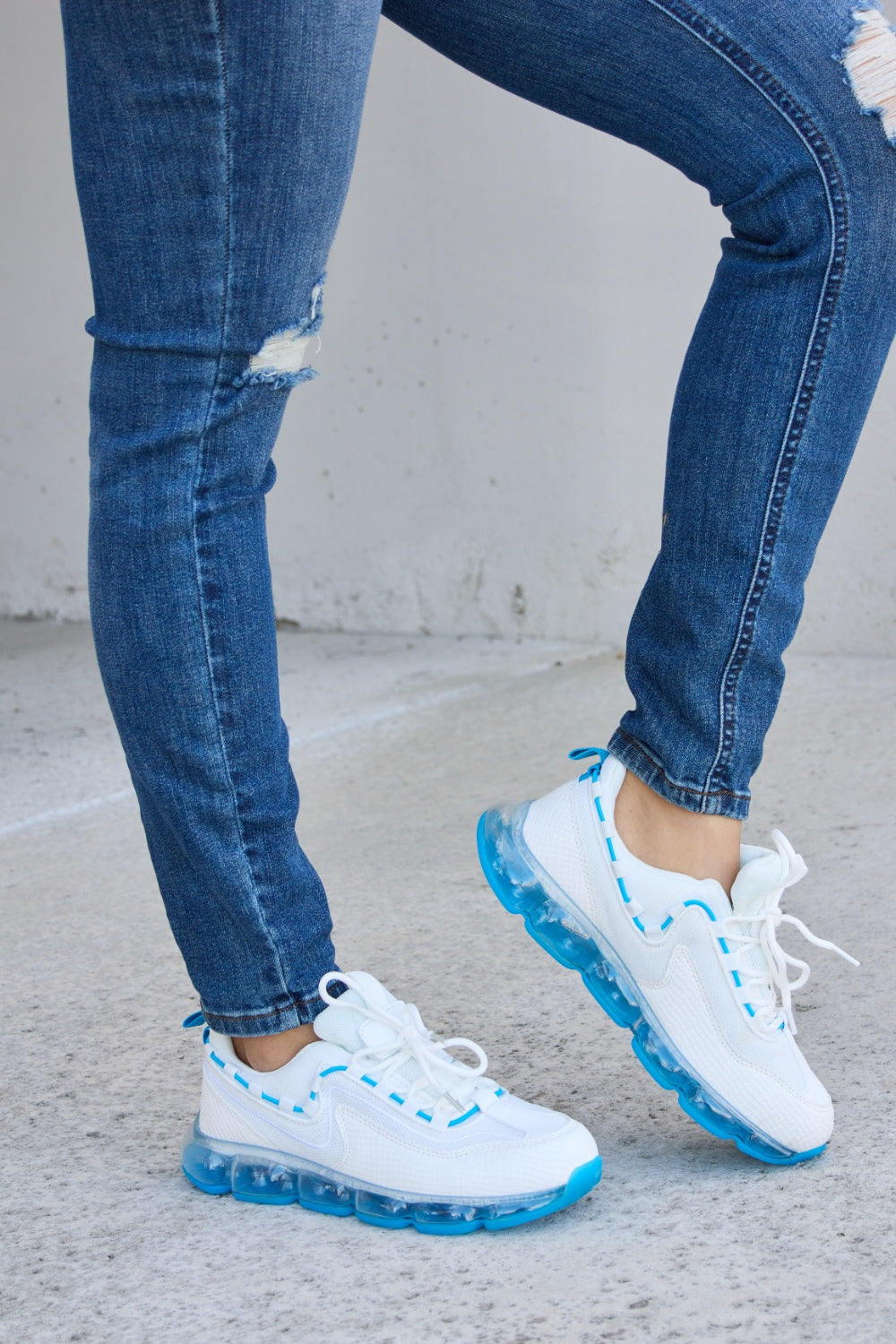 A person wearing ripped jeans and stylish Forever Link Lace-Up Air-Cushioned Athletic Shoes in white with blue accents stands confidently on a concrete surface.