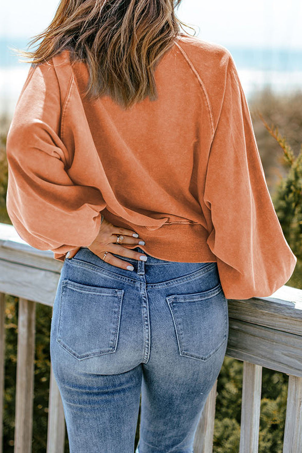 A person wearing an orange washed snap buttons lantern sleeve pullover sweatshirt and blue jeans stands with one hand on their hip, facing away from the camera, leaning slightly on a wooden rail outdoors.