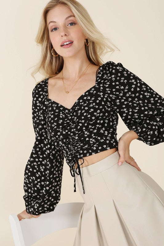 A person with long blonde hair wearing a black ruched floral print crop top with puff sleeves and a beige skirt poses with one hand near their head, looking off to the side.