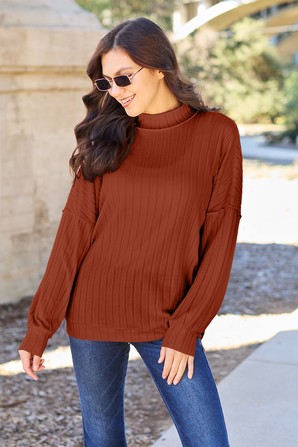 A woman with long hair is wearing a green Basic Bae Full Size Ribbed Exposed Seam Mock Neck Knit Top, blue jeans, and sunglasses. She is standing outdoors and smiling.