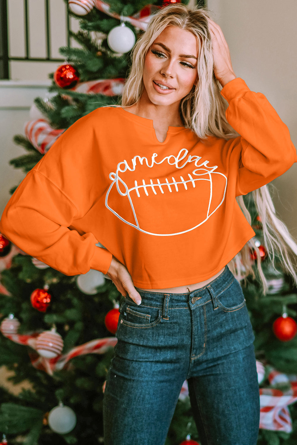 A woman with blonde hair, wearing an Orange Game Day Lettering Rugby Football Notched Neck Sweatshirt and blue jeans, stands next to a decorated Christmas tree.