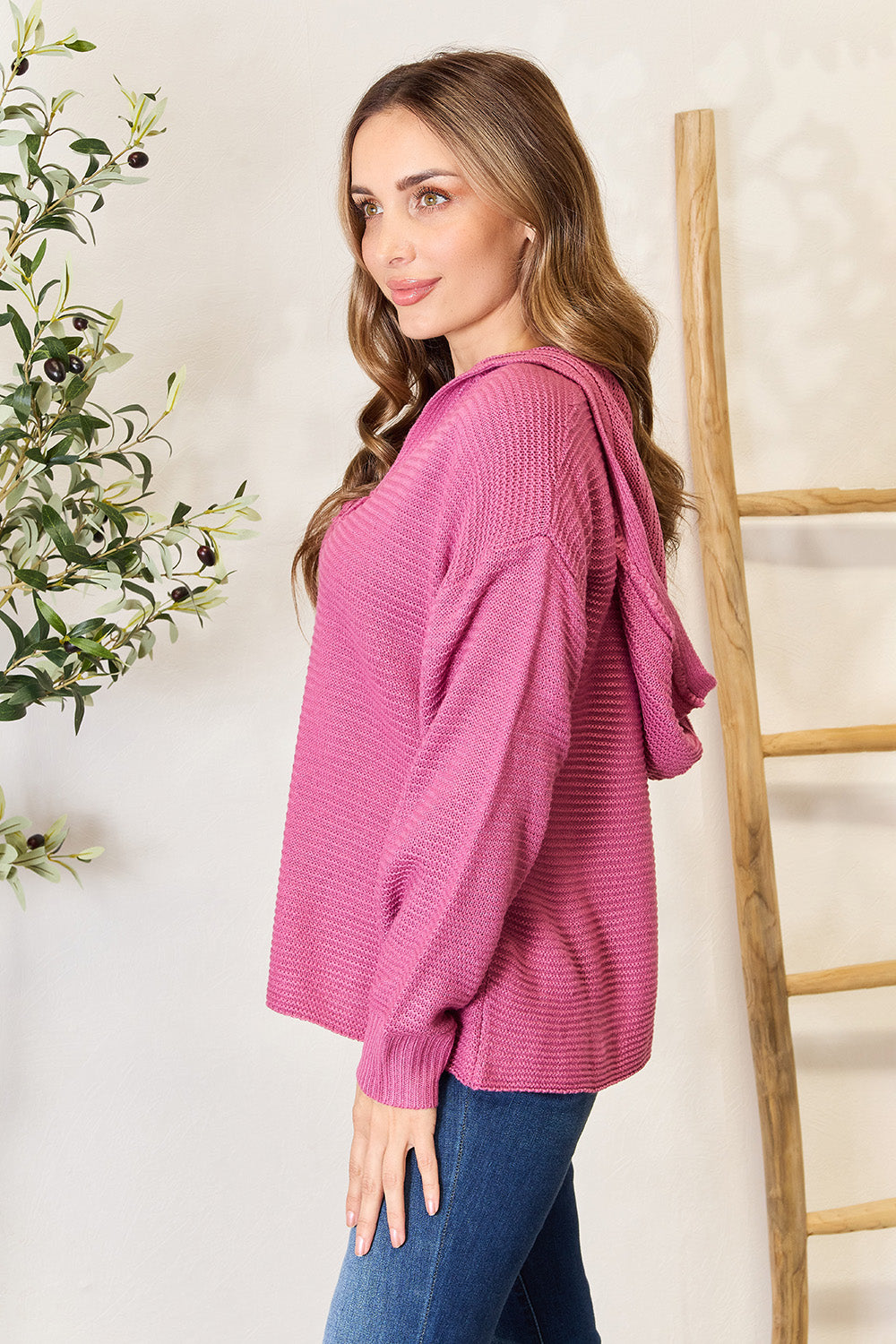 A woman in a stylish Shiny Ribbed Fringed V-Neck Hooded Sweater and jeans stands beside a wooden ladder and a plant.