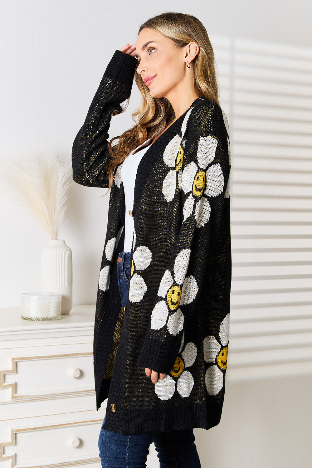 A woman stands indoors wearing the Perfee Floral Button Down Longline Cardigan, which features stylish black fabric adorned with white and yellow smiley face flowers. She pairs it with a white top and blue jeans, looking off to the side while smiling.