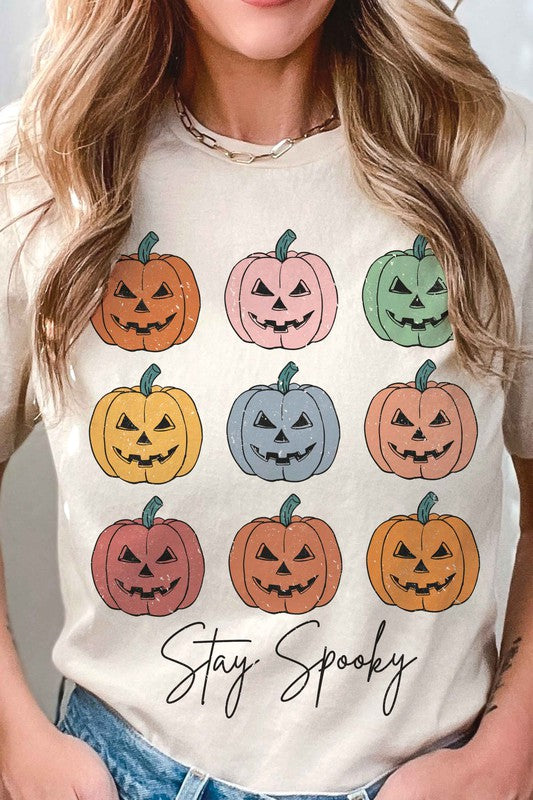 A person wears the STAY SPOOKY MULTI PUMPKINS Graphic Tee in unisex sizing, showcasing nine colorful jack-o'-lanterns and "Stay Spooky" text in a white hue, while holding a drink. They also sport checkered shorts.