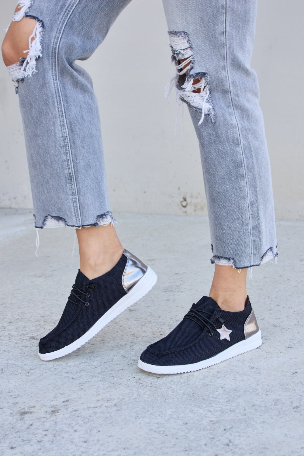 A person stands on a concrete surface, wearing distressed grey jeans and stylish Forever Link Star Lace-Up Flat Sneakers featuring black uppers and white soles.