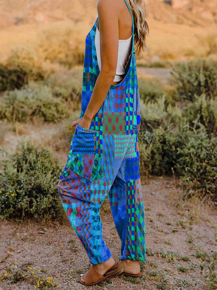 A woman stands outdoors in front of a rocky landscape, wearing the Full Size Printed V-Neck Sleeveless Jumpsuit and a brown brimmed hat. She is smiling and holding the brim of her hat with one hand, exuding the carefree vibe of this vibrant and colorful jumpsuit.