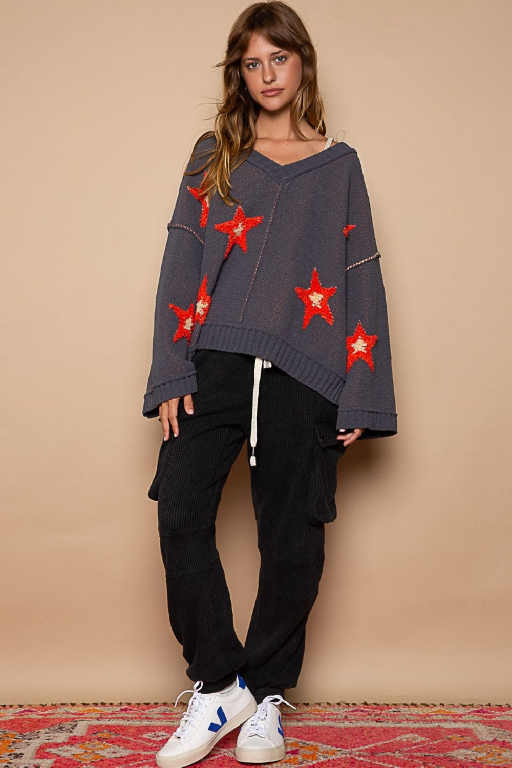 A person with long hair wears a stylish and cozy POL Long Sleeve Star Patch Sweater adorned with color-blocked star patterns in red, paired with black pants, standing against a tan background.