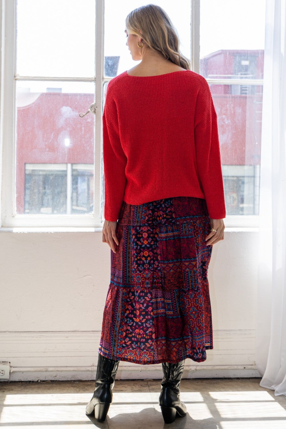 A person stands near a window exuding bohemian charm in a red sweater, an ODDI Full Size Printed Tiered Maxi Skirt, and black boots.