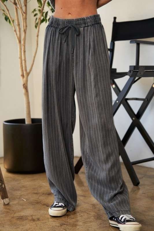 A person wears a casual outfit called the Stripe Button Down Shirt and Long Pants Set, which features a loose-fitting pinstriped design, paired with sneakers. They stand indoors near a potted plant and a director's chair.