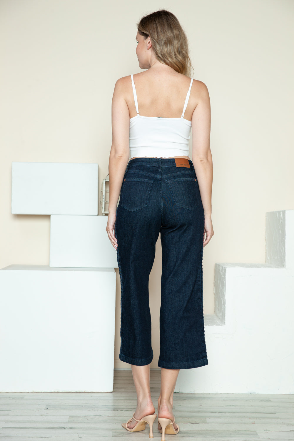 Person wearing Judy Blue Full Size Side Seam Braid Detail Crop Wide Leg Jeans and beige strappy heels, standing on a wooden floor with a white background.