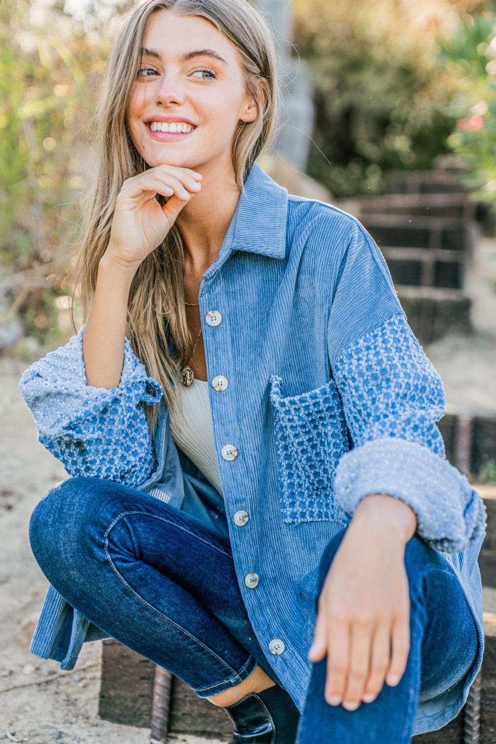 A woman stands outdoors wearing the And The Why Button Down Corduroy Contrast Texture Long Sleeve Shacket over a white top and blue jeans. She has long hair and wears a belt with a prominent buckle.