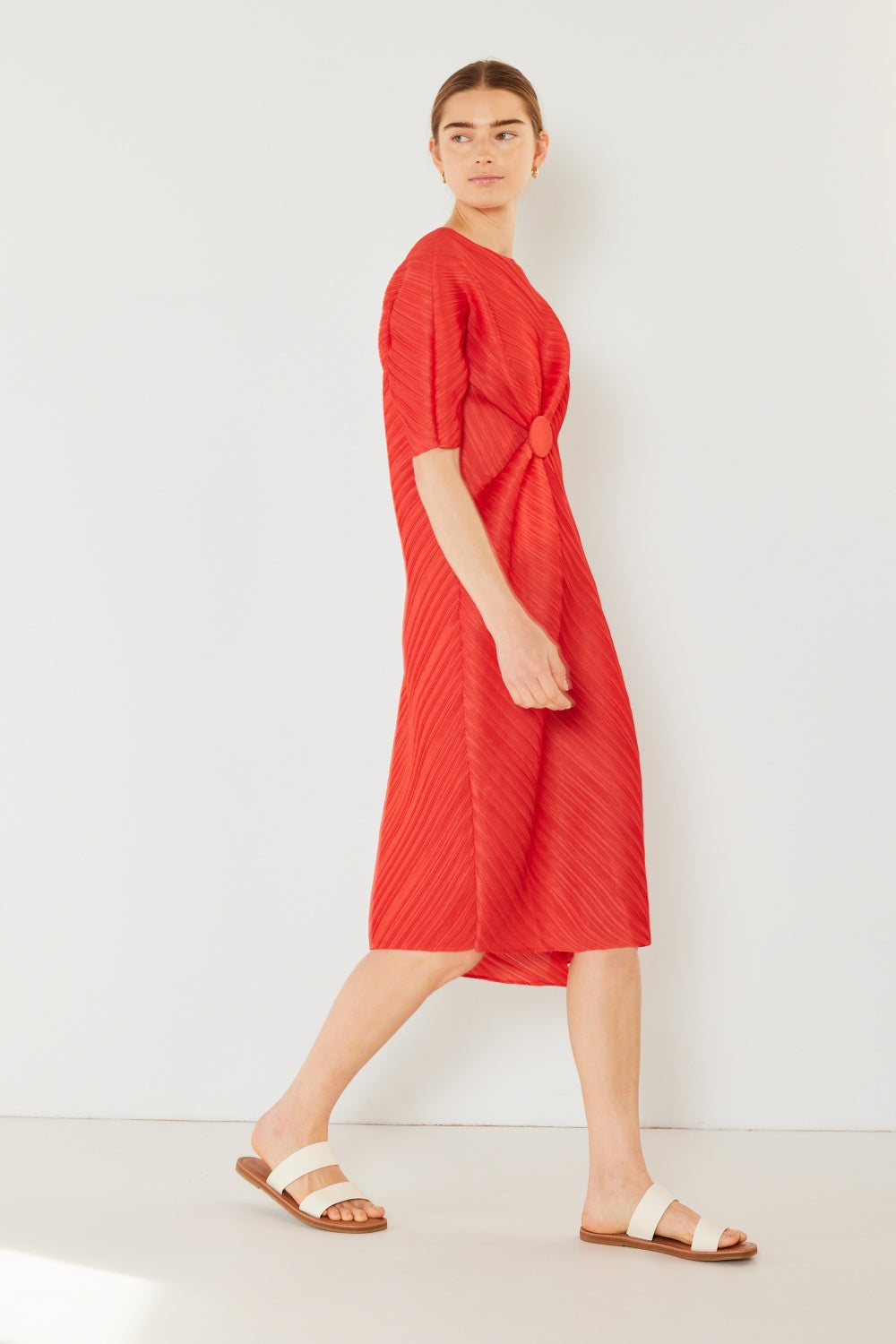 A woman stands against a plain backdrop wearing the stylish Marina West Swim Pleated Dolman Sleeve Dress, which features a knee-length, short-sleeved design in a textured brown fabric with an eye-catching circular detail at the waist. She completes her look with white sandals. This modern chic dress exudes versatility, making it perfect for various occasions.