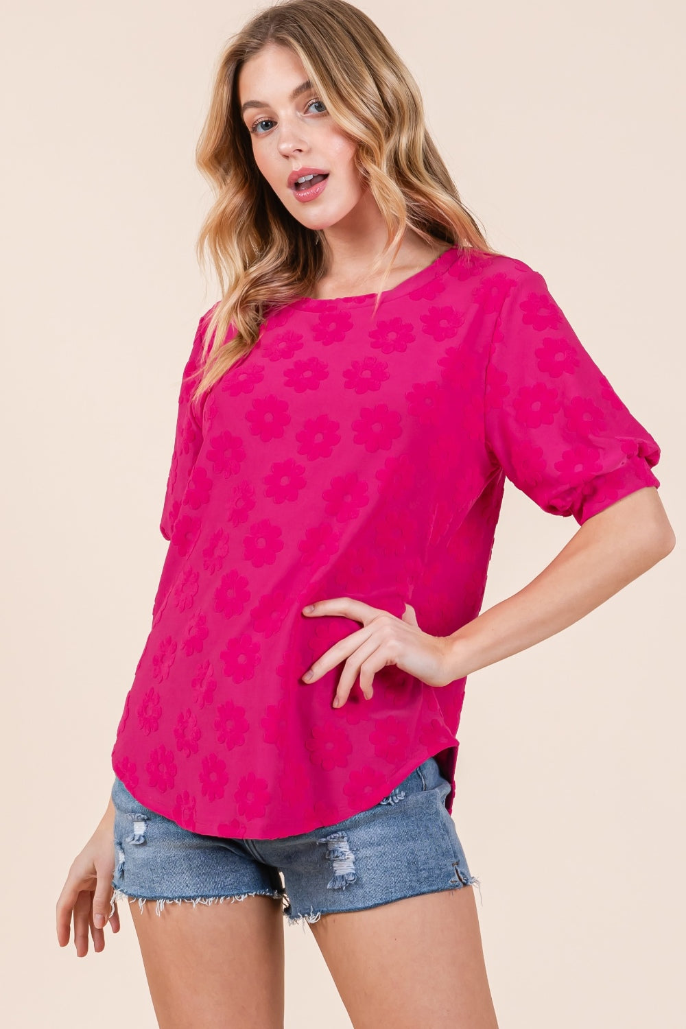 A woman showcases her versatile style in the BOMBOM Textured Floral Pattern Top, with its bright pink hue and feminine design, paired effortlessly with denim shorts against a plain background.