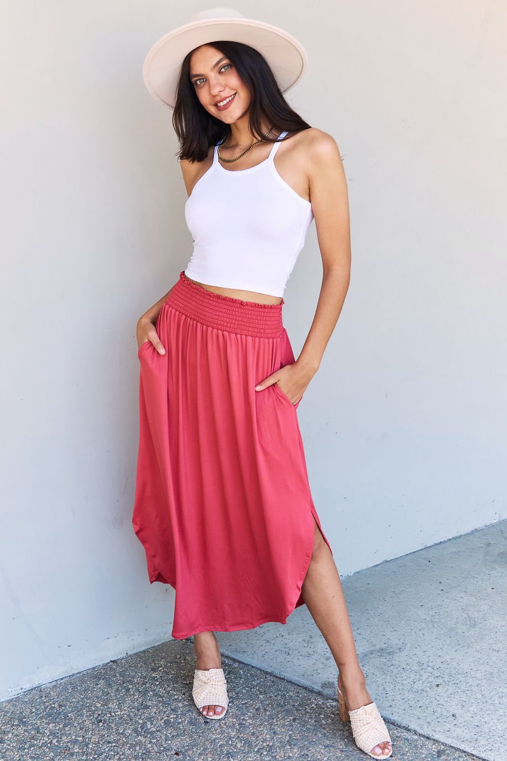 A person wearing a white top and the Doublju Comfort Princess Full Size High Waist Scoop Hem Maxi Skirt in an elegant coral color, paired with tan sandals, stands in front of a plain wall.