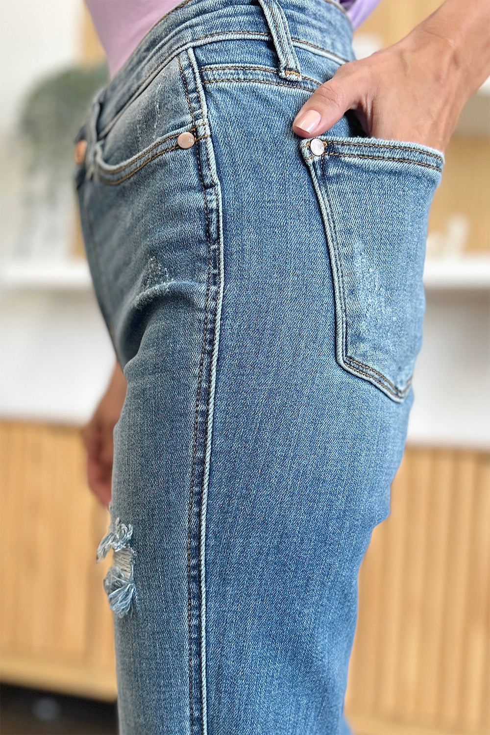 A person wearing Judy Blue Full Size Distressed Raw Hem Bootcut Jeans and a light purple top stands on a polished floor with a wooden cabinet in the background.