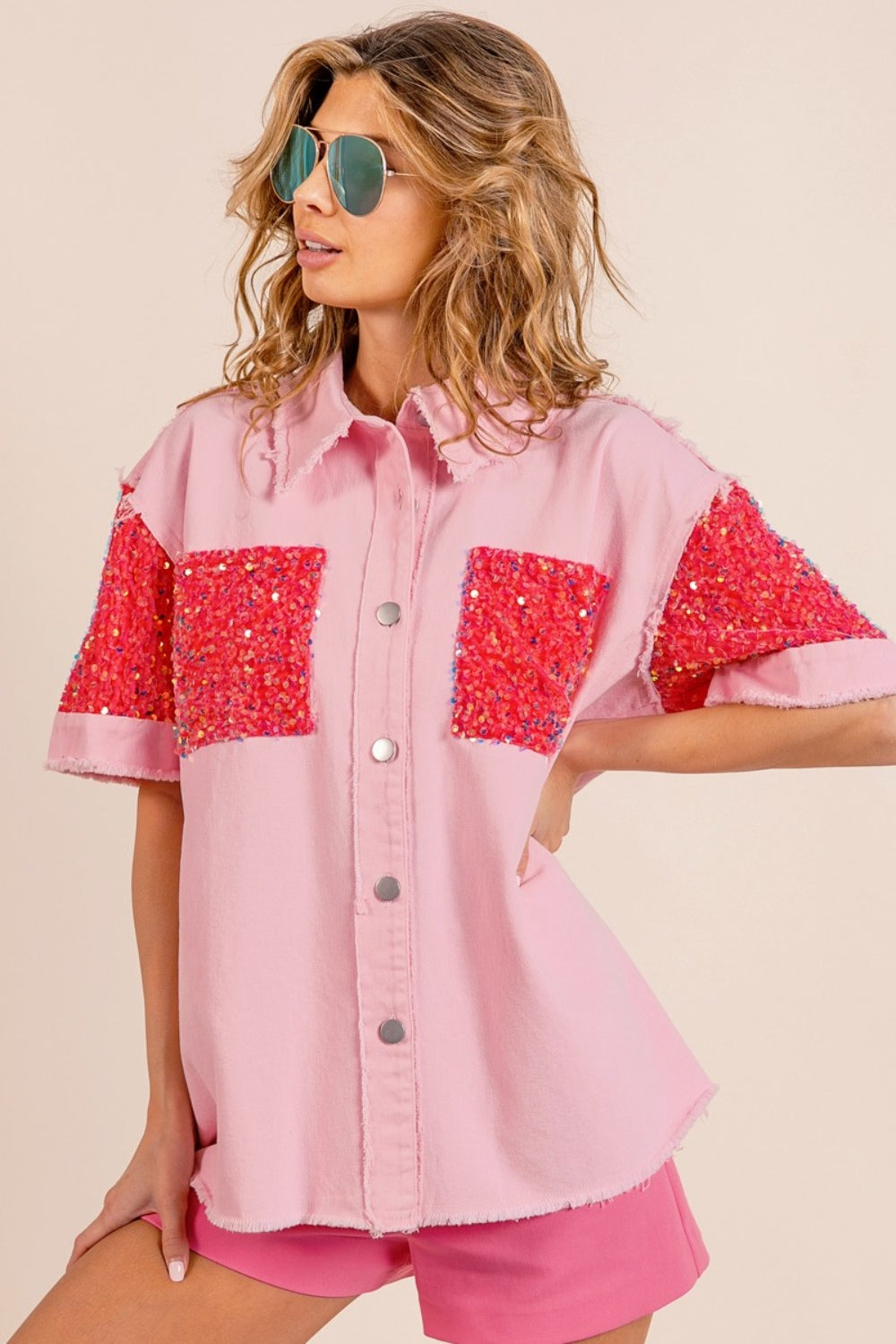 A woman poses in the BiBi Sequin Detail Raw Hem Short Sleeve Shirt, a pink button-up with red sequin detail pockets, paired with matching pink shorts and reflective aviator sunglasses.