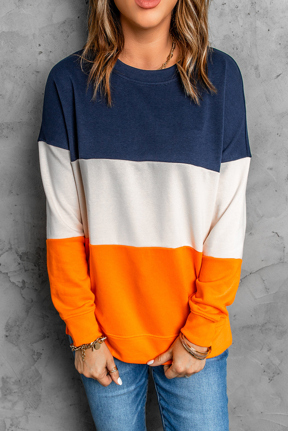 Person with wavy, shoulder-length hair wearing a Colorblock Orange Contrast Stitching Sweatshirt with Slits, paired with blue skinny fit jeans, standing with back to the camera against a gray textured wall.