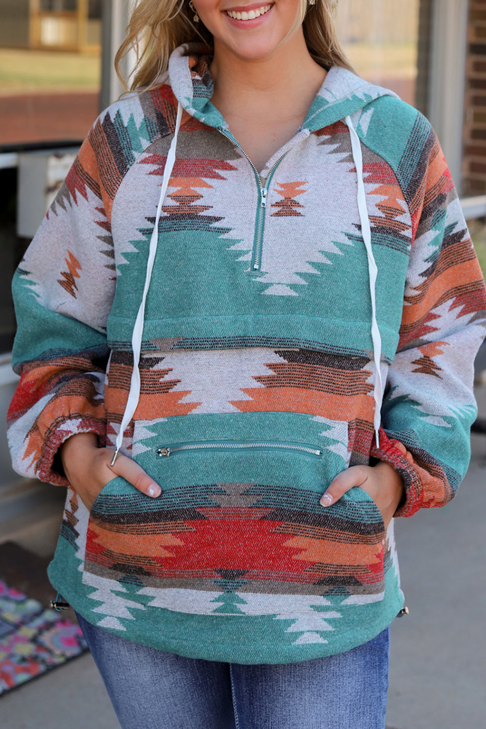 A woman with long blonde hair is standing outside, facing away from the camera. She wears a Multicolor Aztec Print Zipped Split Neck Hoodie and jeans. A rug and potted plant are visible in the background, adding to the cozy vibe of her super comfortable outfit.