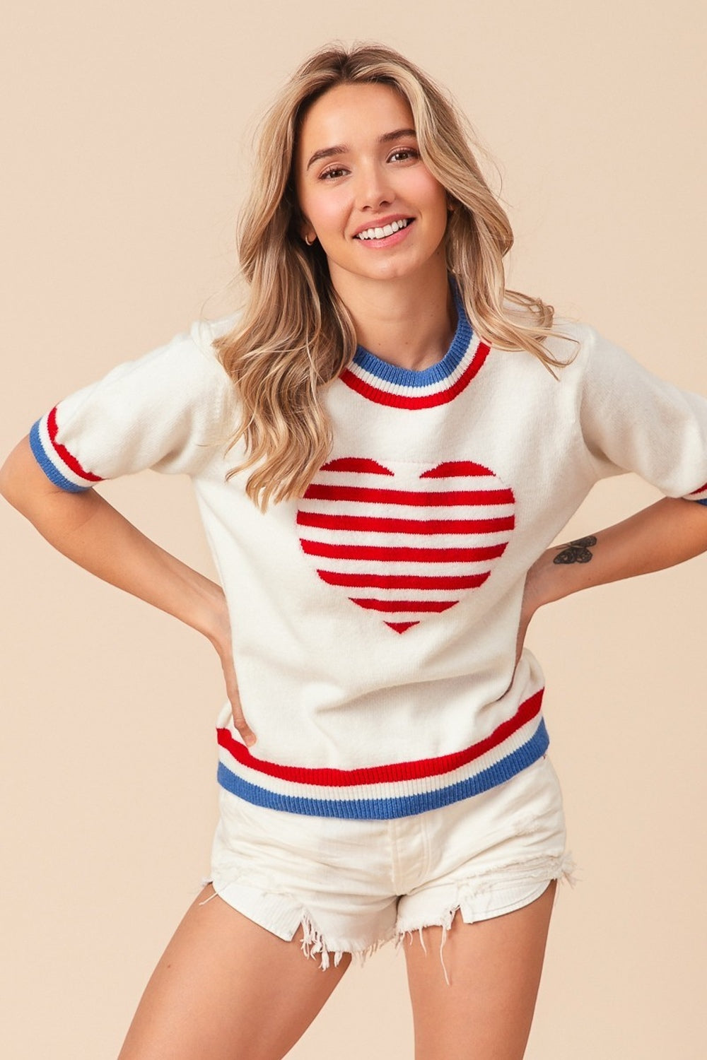 A person wearing the BiBi US Flag Theme Striped Heart Sweater, featuring a white short-sleeve design with red and blue stripes resembling the US flag and adorned with a red heart motif. They are also wearing white shorts and smiling with hands on hips.