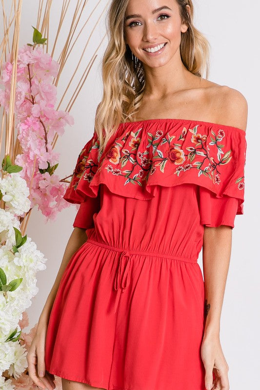 Wearing the stylish Floral Embroidered Off Shoulder Romper, a woman poses gracefully, showcasing its ruffle detail and making it an ideal addition to any summer wardrobe.