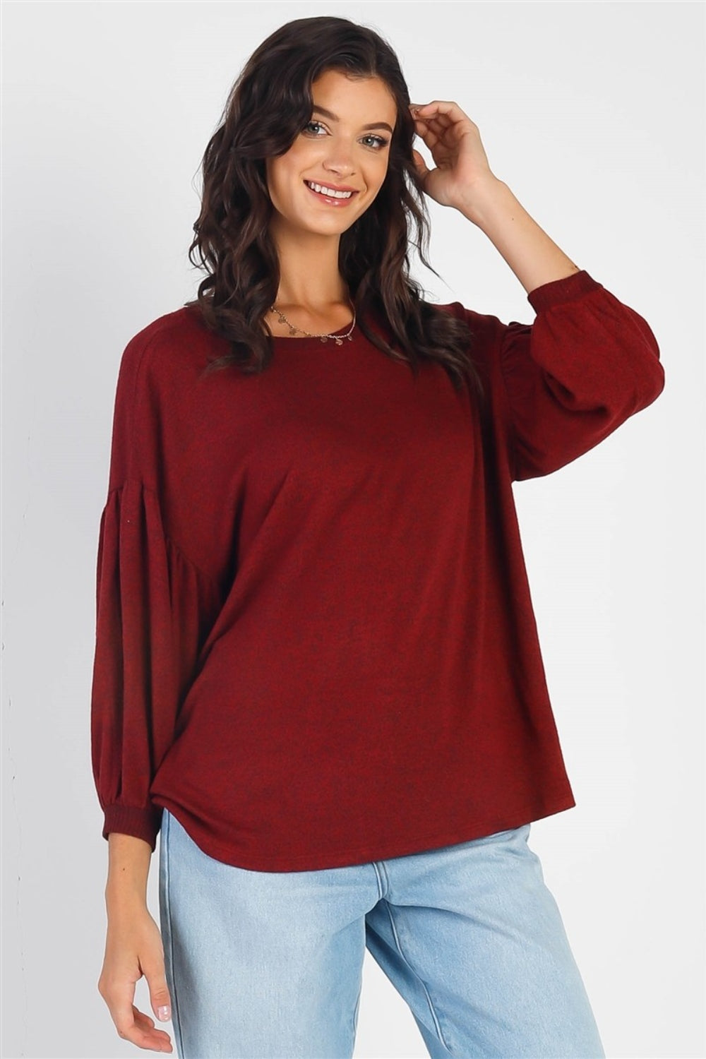 A person with long dark hair stands against a white background, smiling and raising one hand while wearing the trendy and stylish Cherish Apparel Drop Shoulder Puff Sleeve Top in burgundy paired with light blue jeans.