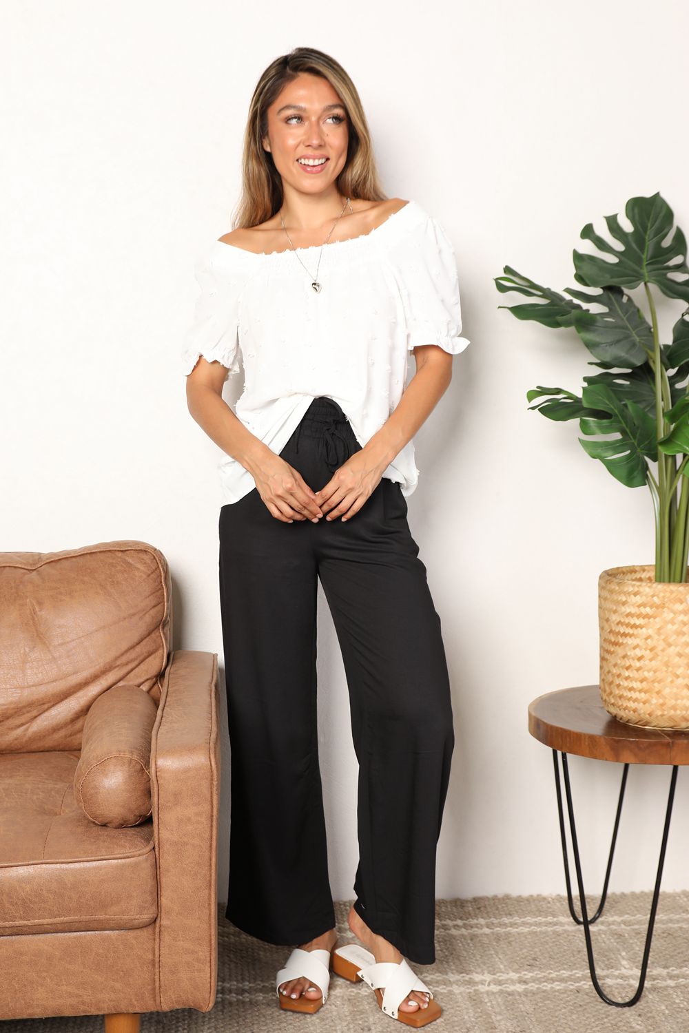 A person wearing the Mandy Swiss Dot Puff Sleeve Square Neck Blouse in white, paired with black pants, stands in front of a light background with green leaves visible on the right side.