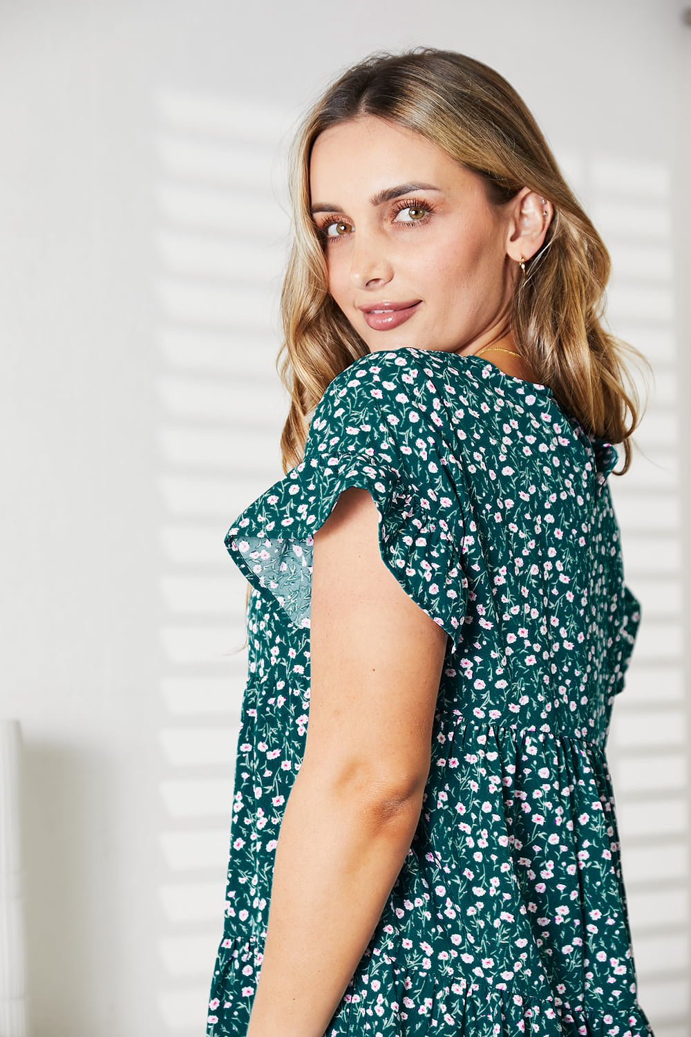 A woman wearing the Double Take Short Flounce Sleeve Tiered Dress in pink and red leopard print, complete with a ruffled hem and short flounce sleeves, smiles as she adjusts her brown hat. She stands near a plant, embodying the perfect boho-chic look.