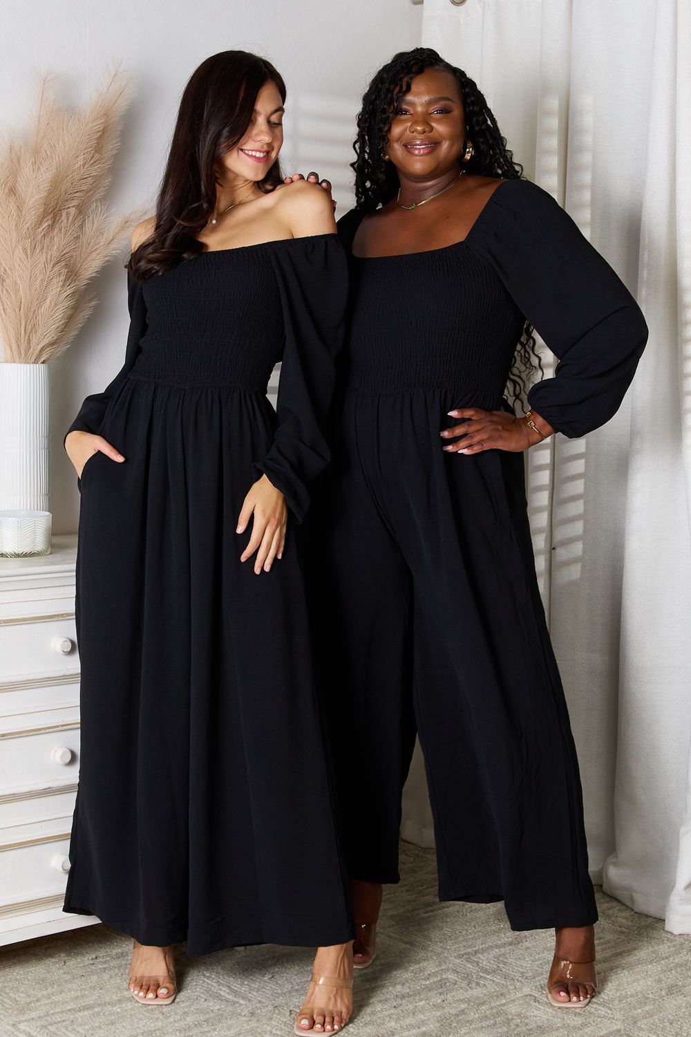 Two women in Double Take Square Neck Jumpsuits with pockets stand side by side in a room decorated with white accents and pampas grass in a vase. Both are smiling, have their hands in their pockets, and are wearing high heels.